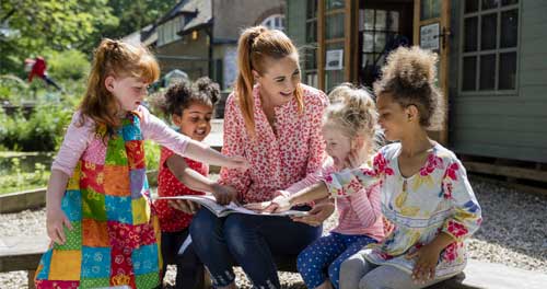 A front view of a childcare worker and some of her young female pupils. They are storytelling in a story circle and are having fun listening as she reads and encourages them to join in.